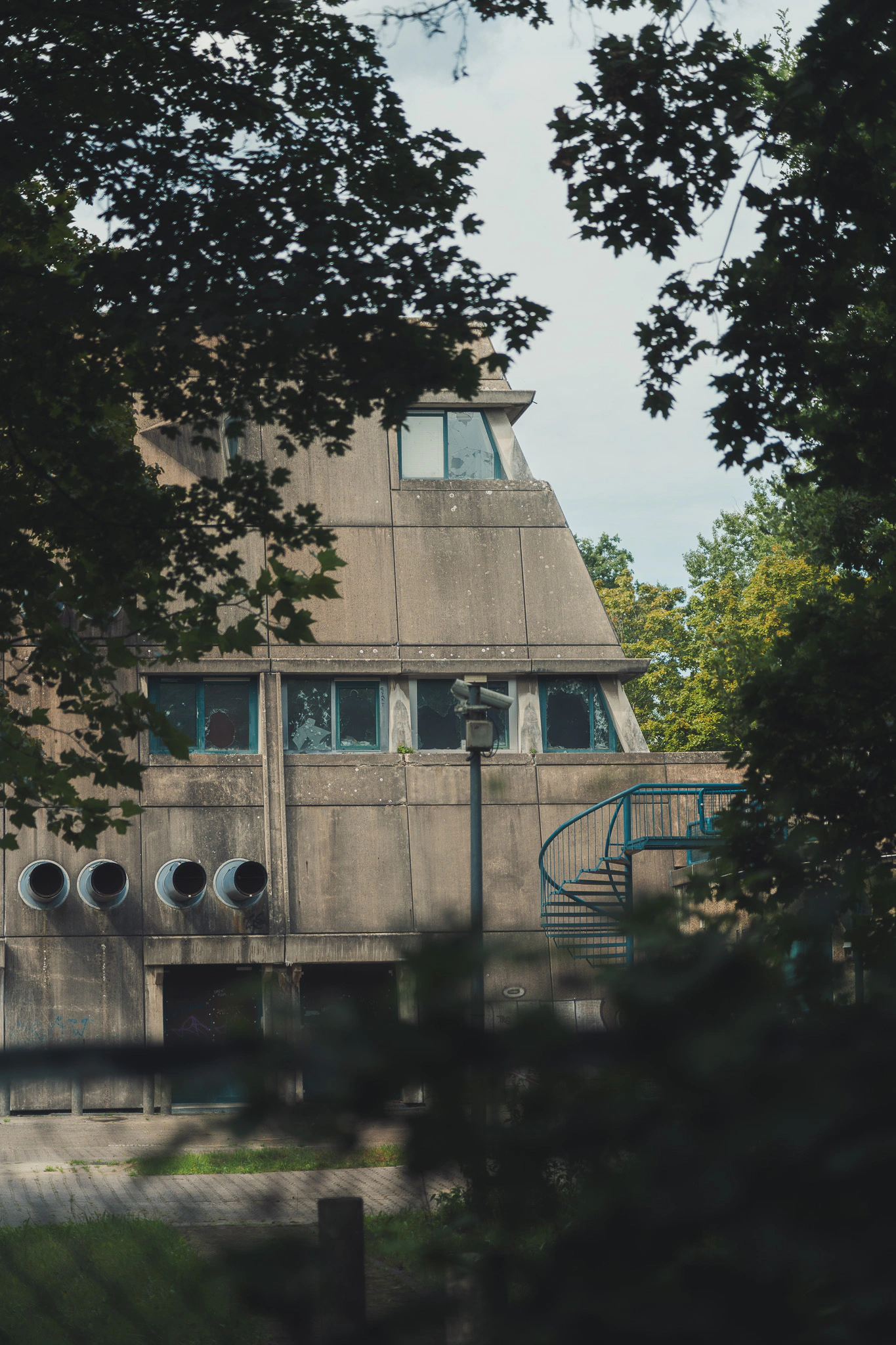 Sloped facade of the Mäusebunker with blue ventilation pipes, seen through trees