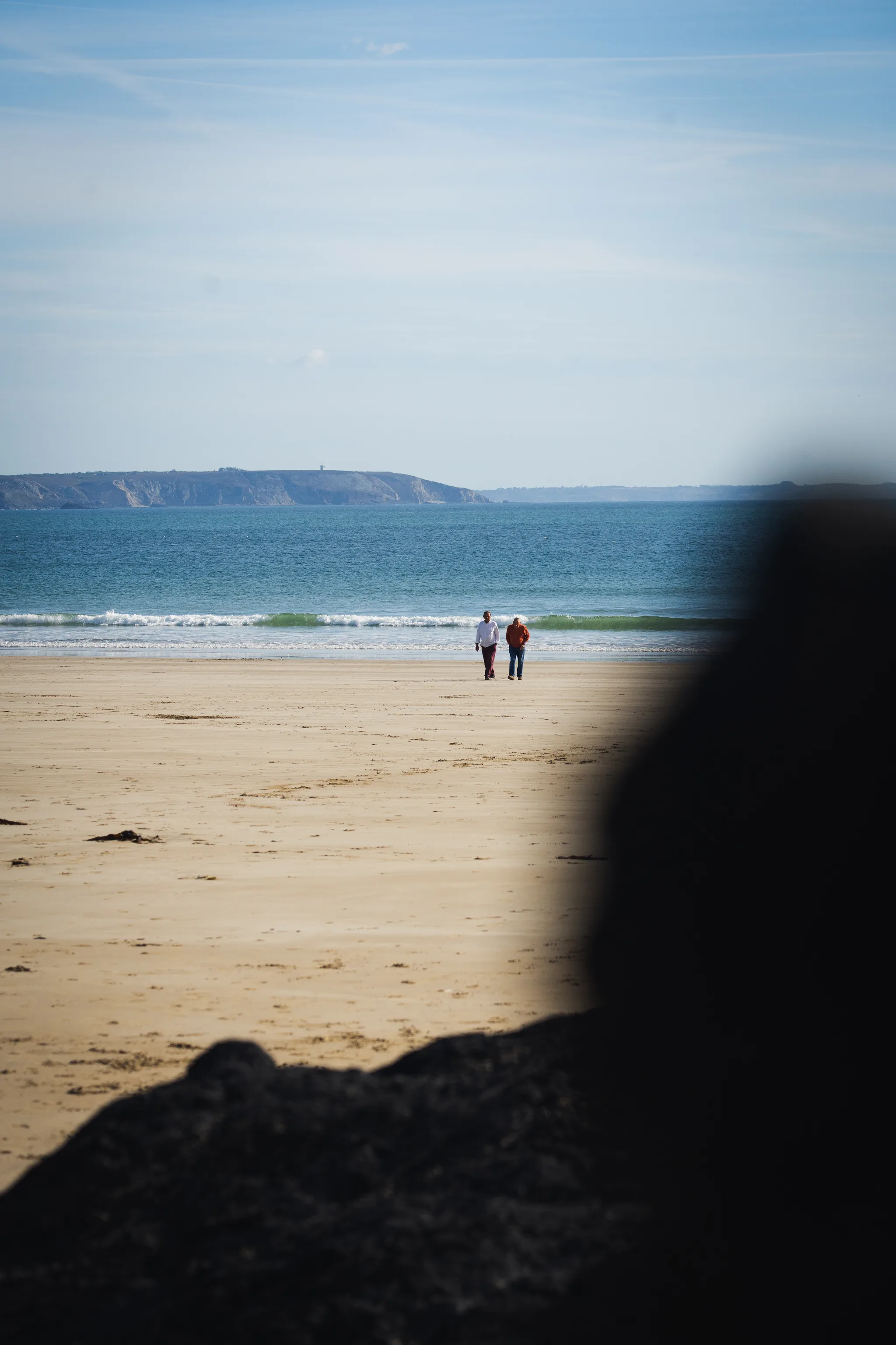Two people on the beach seen past a dark rock in the foreground