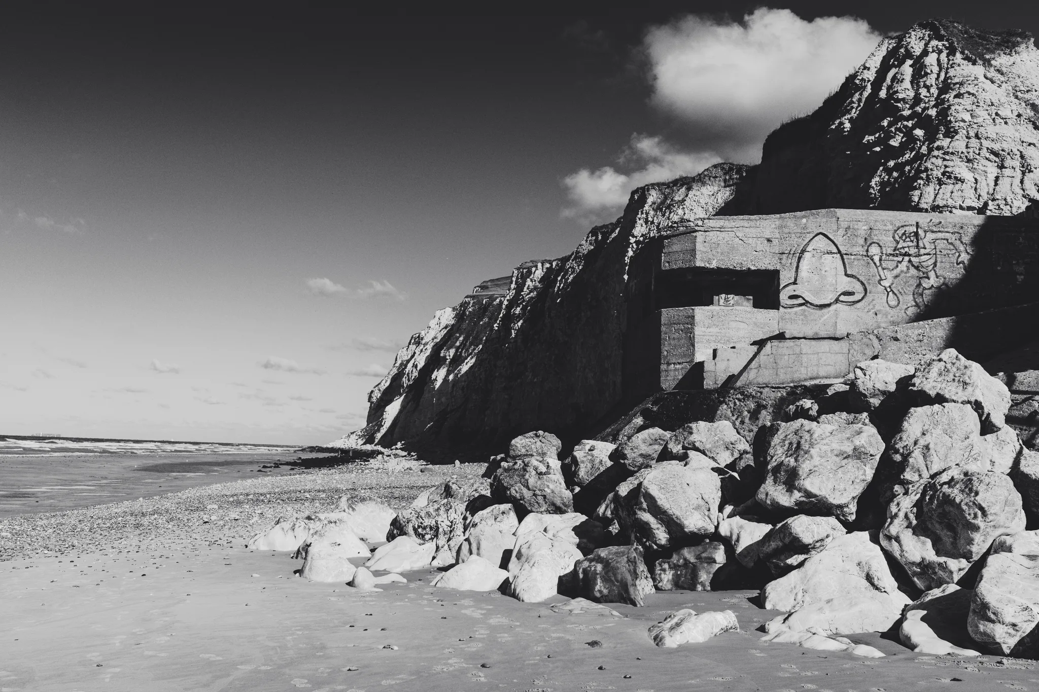 Concrete bunker with graffiti set into a chalk cliff above boulders on the beach, black and white