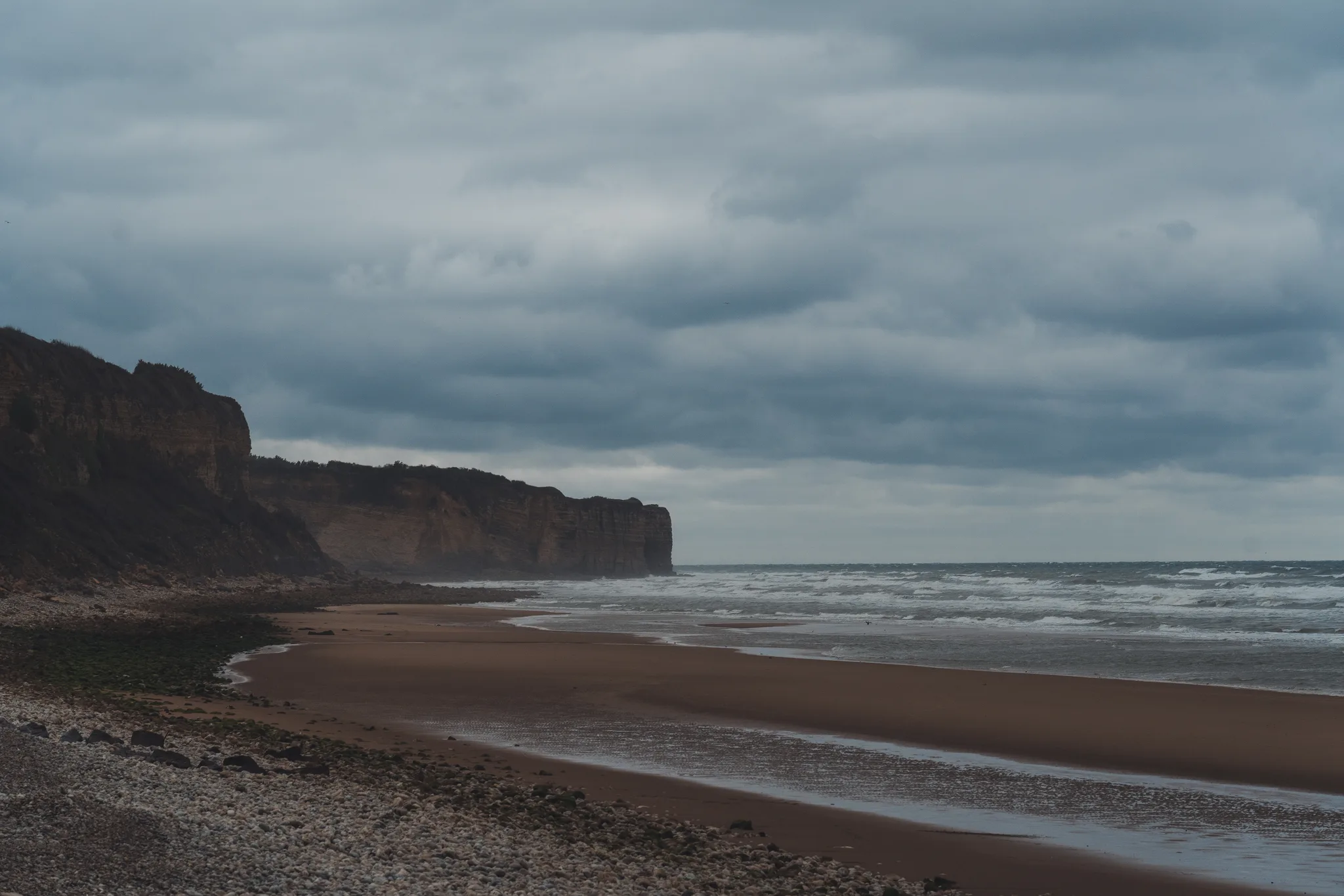 Reddish cliffs above a wide beach with surf under dark clouds