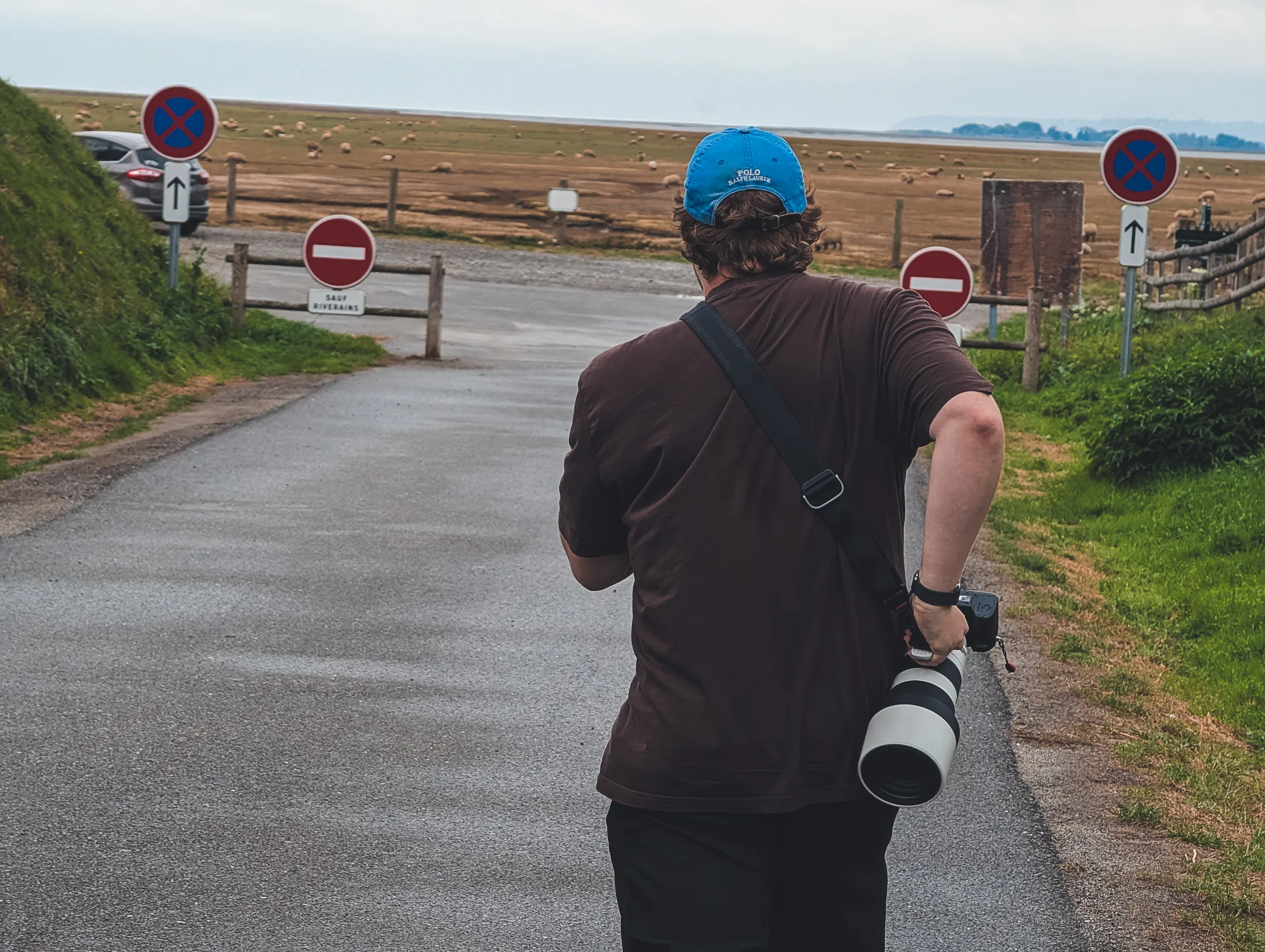 Man in a blue cap carrying a camera with a large telephoto lens down a wet road