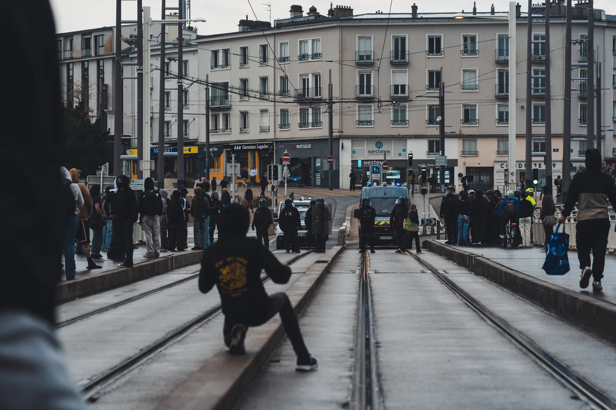 Crowd of hooded people on the tram tracks, a police van at the square beyond