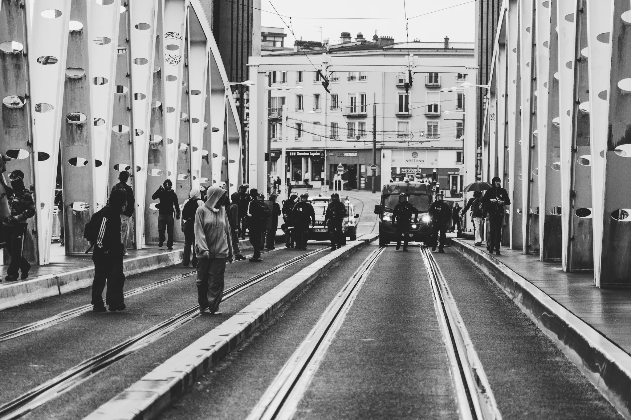 People walking along the tram tracks on the bridge, a police van in the distance, black and white