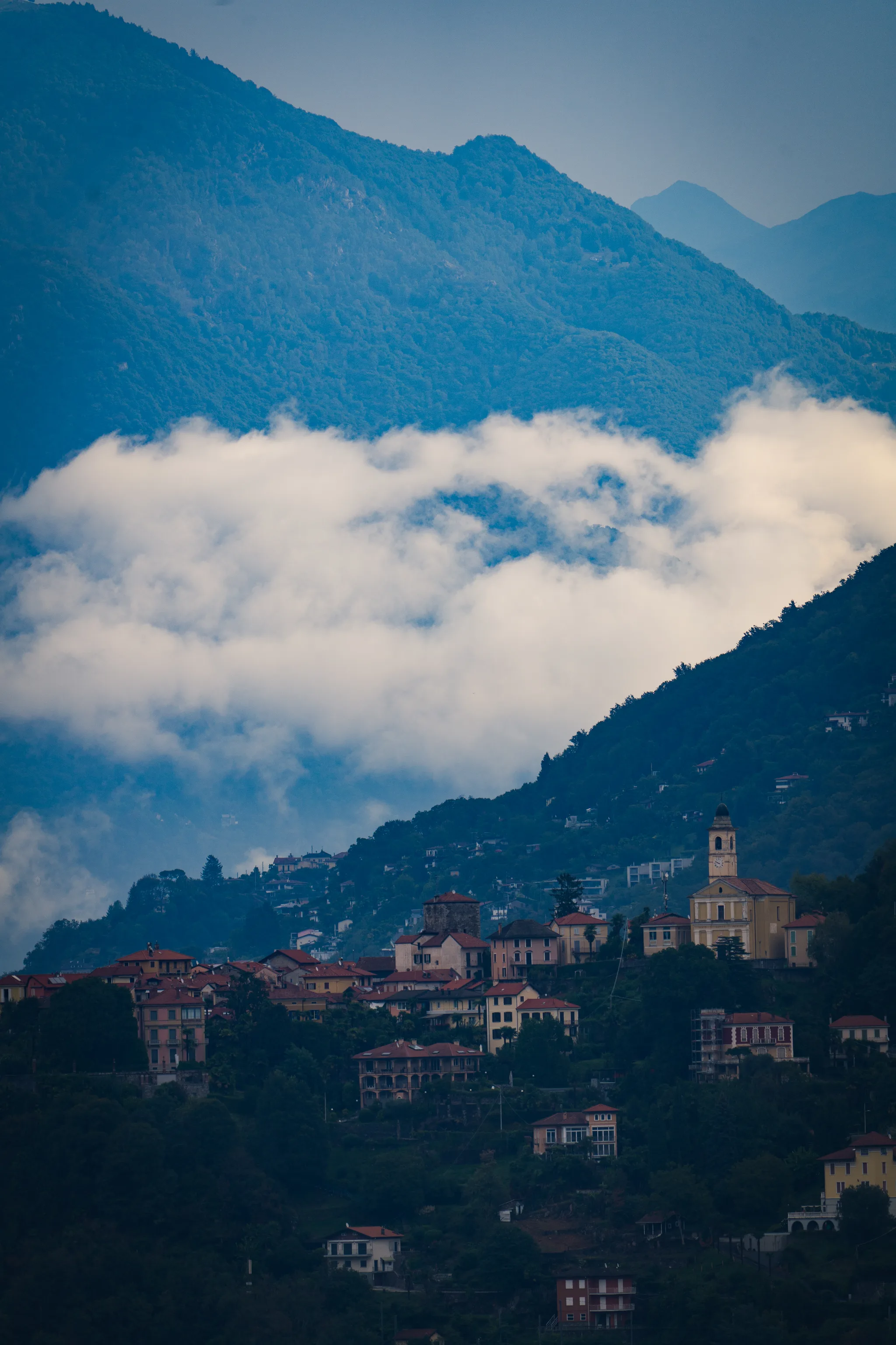 Hillside village with a bell tower below a band of low cloud lying across the mountain
