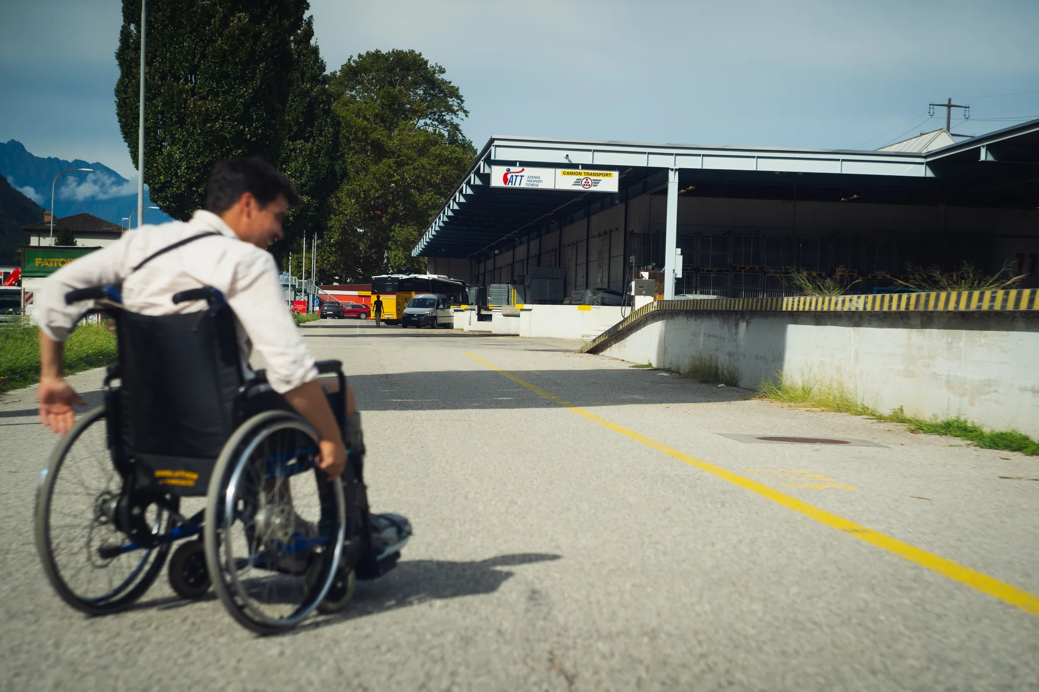 Man in a wheelchair rolling across an empty yard towards a truck depot, mountains in the background