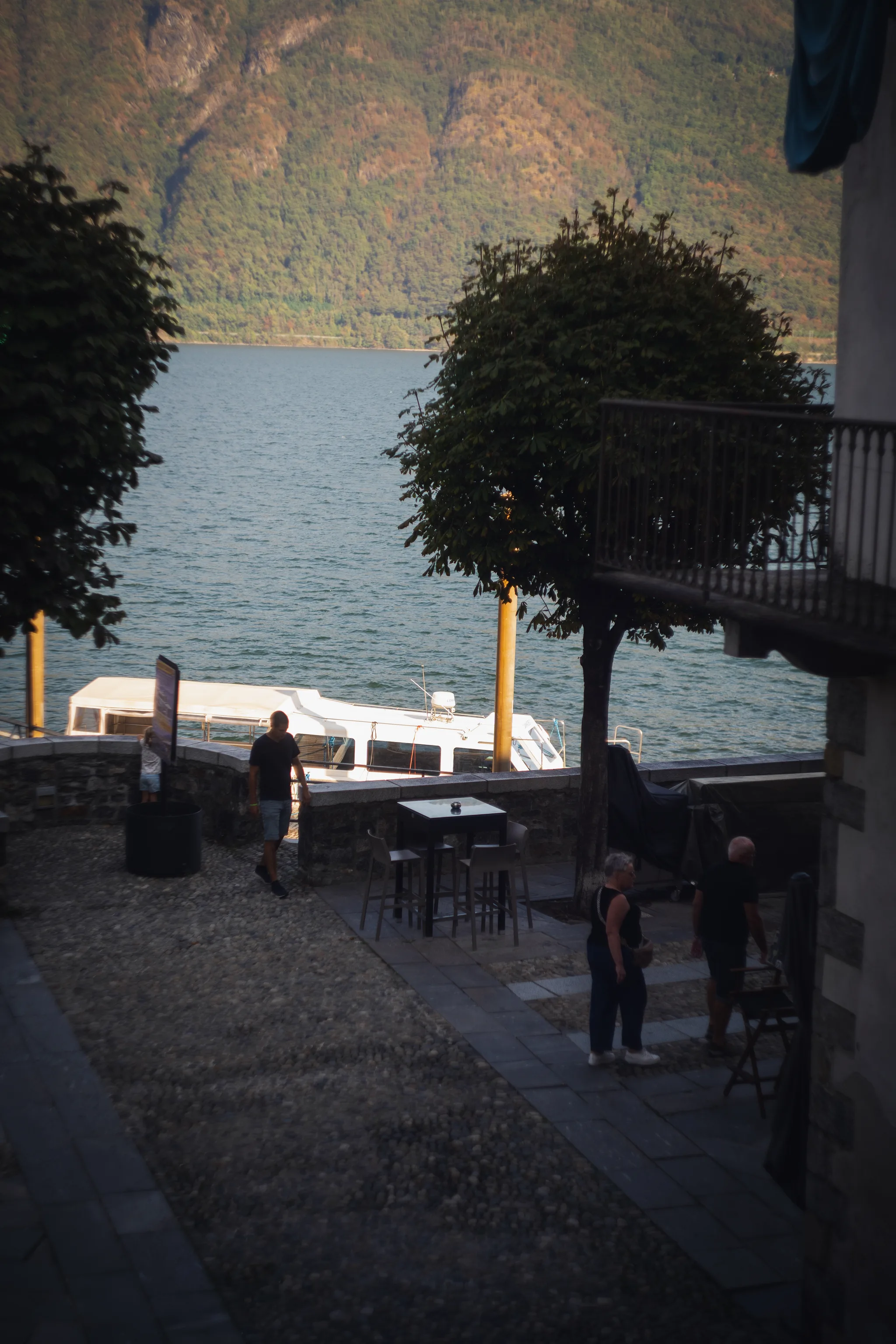 Lakefront terrace with tables, a moored boat and people walking, the lake behind
