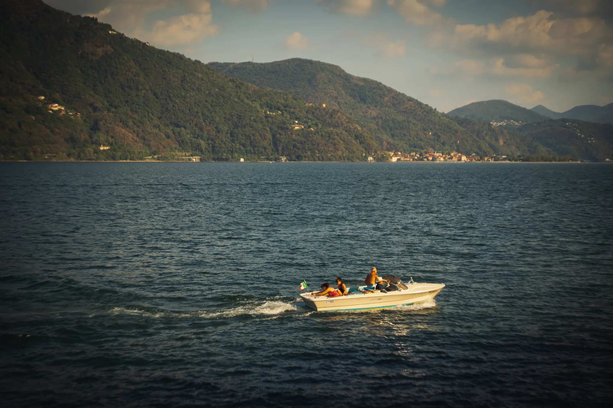 Small motorboat with an Italian flag crossing the lake in late light, hills and a village behind