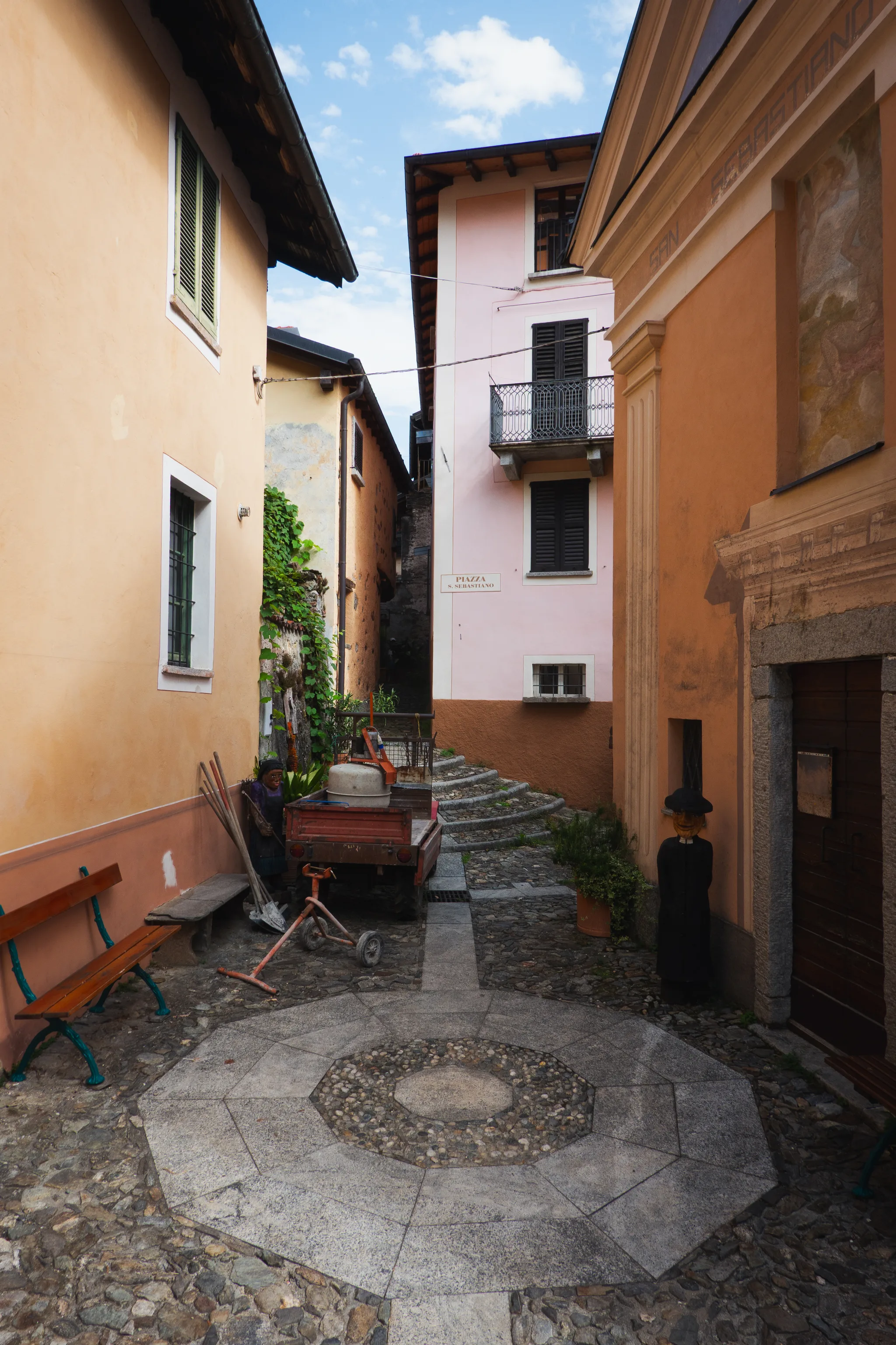 Small square with an octagonal stone inlay, a wooden cart and a dressed-up figure by a door, Piazza S. Sebastiano sign on a pink house