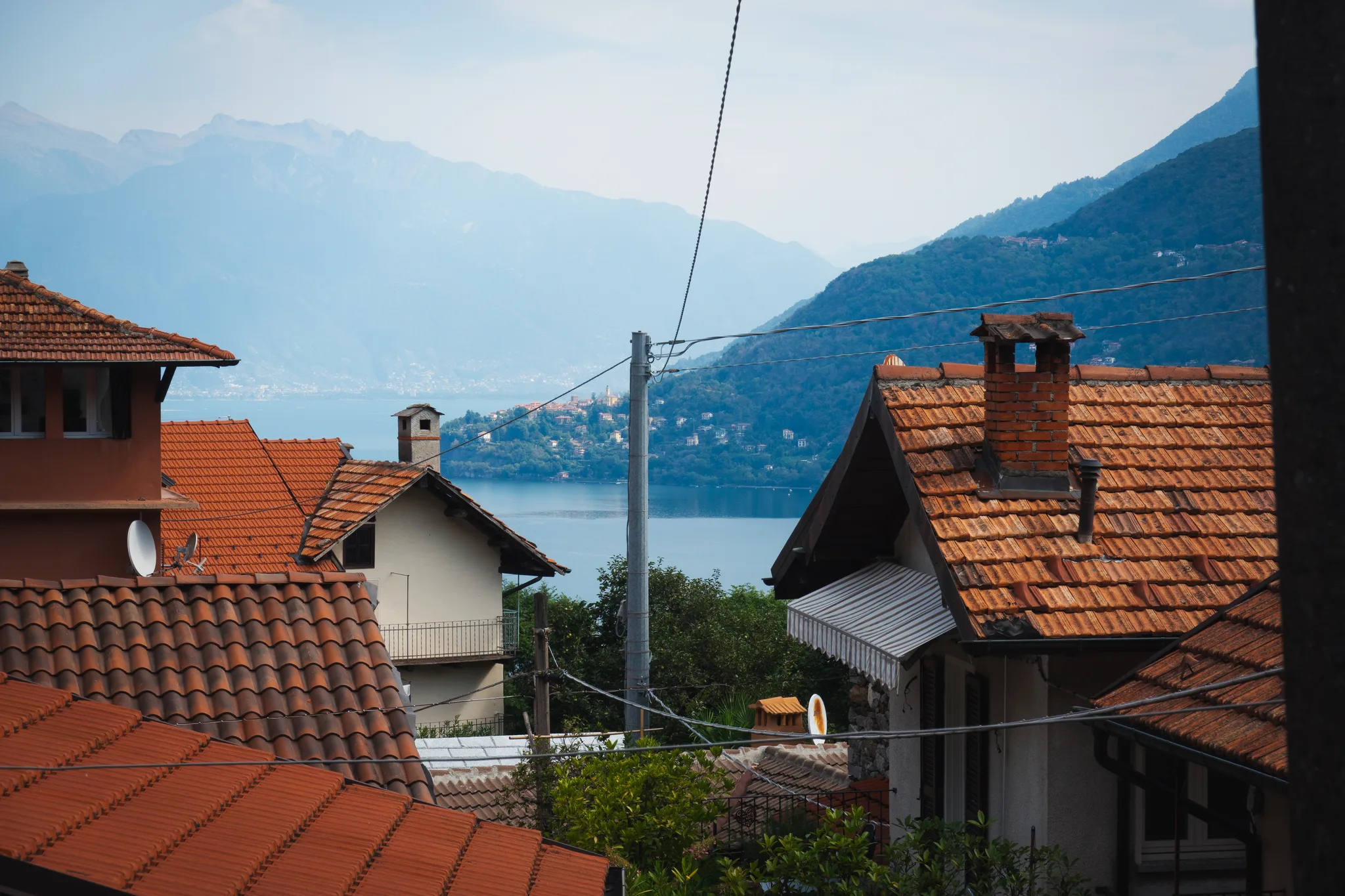 Terracotta roofs and power lines with the lake and a promontory village behind, mountains in haze
