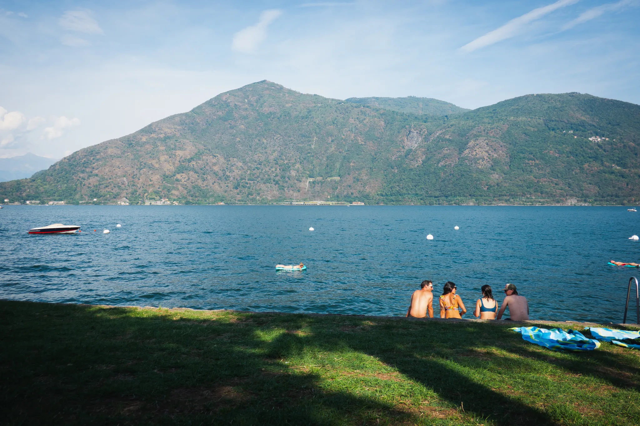 Four swimmers sitting at the edge of a lawn by the lake, a motorboat and buoys on the water, a green mountain across