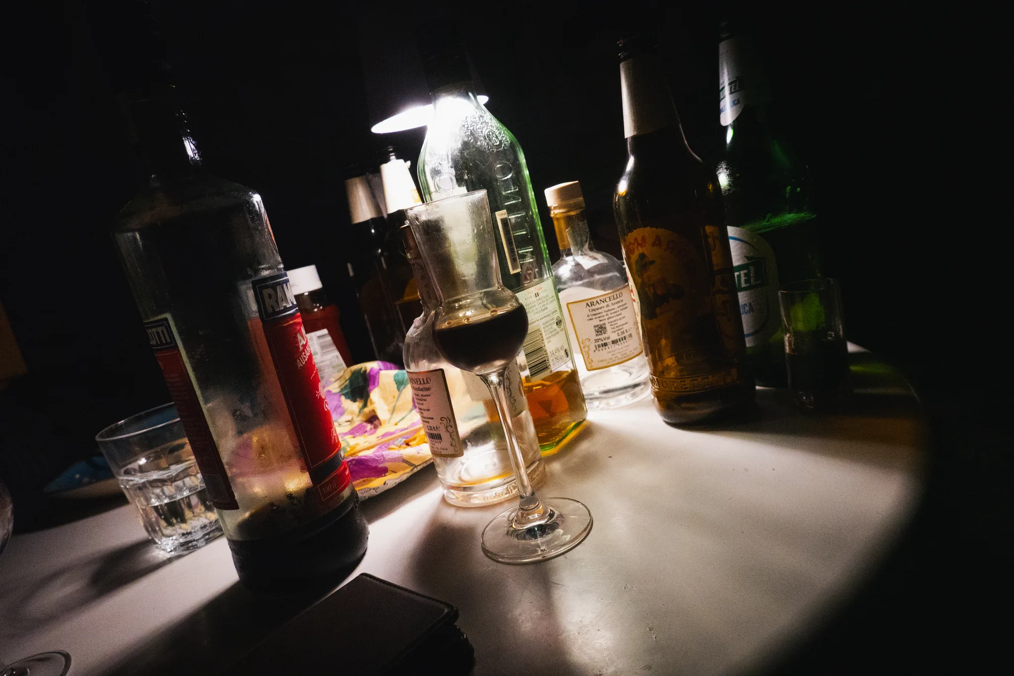 Bottles and a glass of red on a table under a single lamp at night
